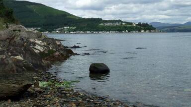 A rocky shoreline with a large boulder in the water, a distant village on the hills, and a cloudy sky.