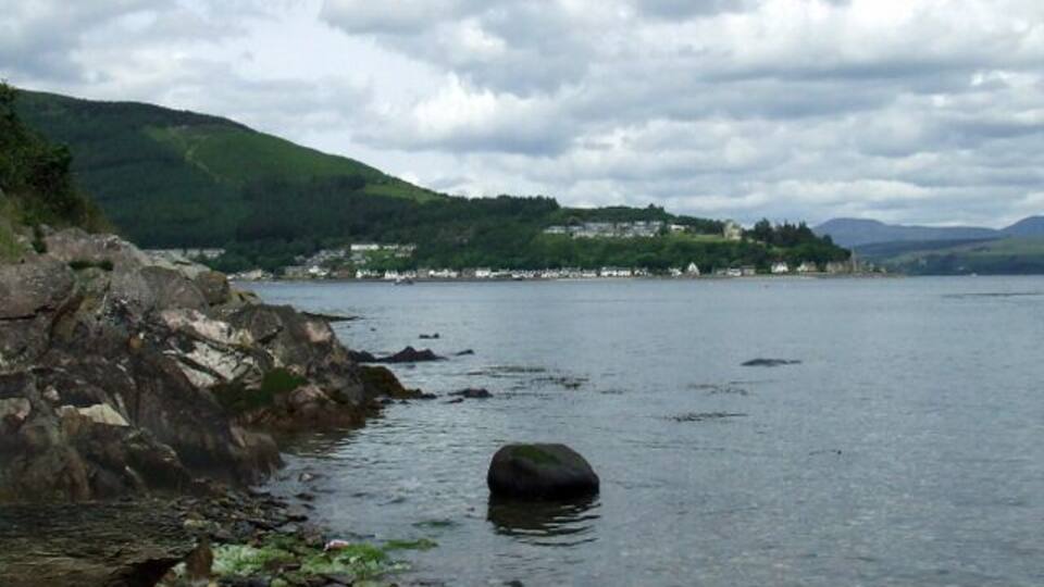 A rocky shoreline with a large boulder in the water, a distant village on the hills, and a cloudy sky.