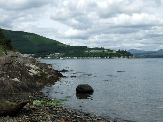 A rocky shoreline with a large boulder in the water, a distant village on the hills, and a cloudy sky.