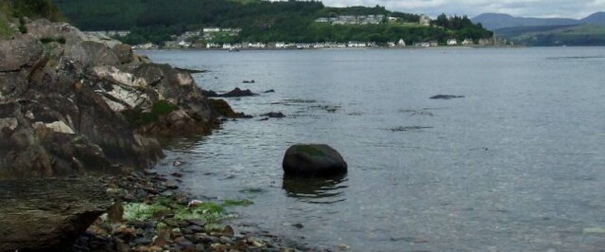 A rocky shoreline with a large boulder in the water, a distant village on the hills, and a cloudy sky.