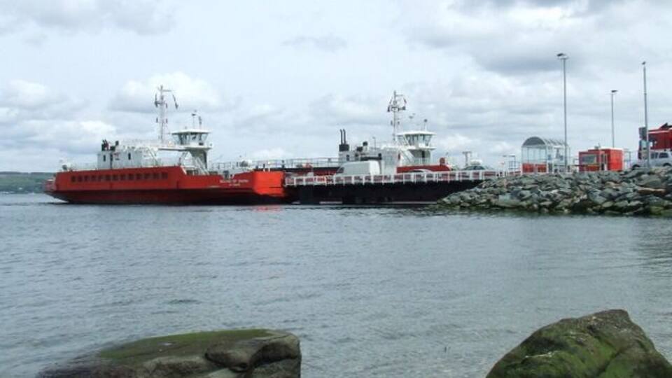 A red ferry docked at a pier with a rocky shoreline and a cloudy sky.