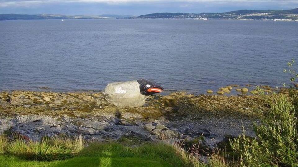 A rocky shoreline with a large boulder and a small orange object on the grass.