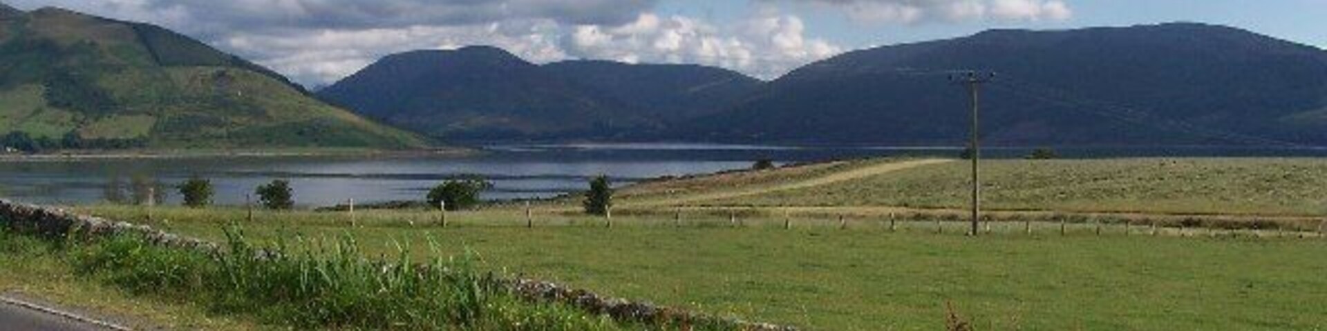 Paisagem rural com estrada, campos gramados, um corpo d'água e montanhas ao fundo.
