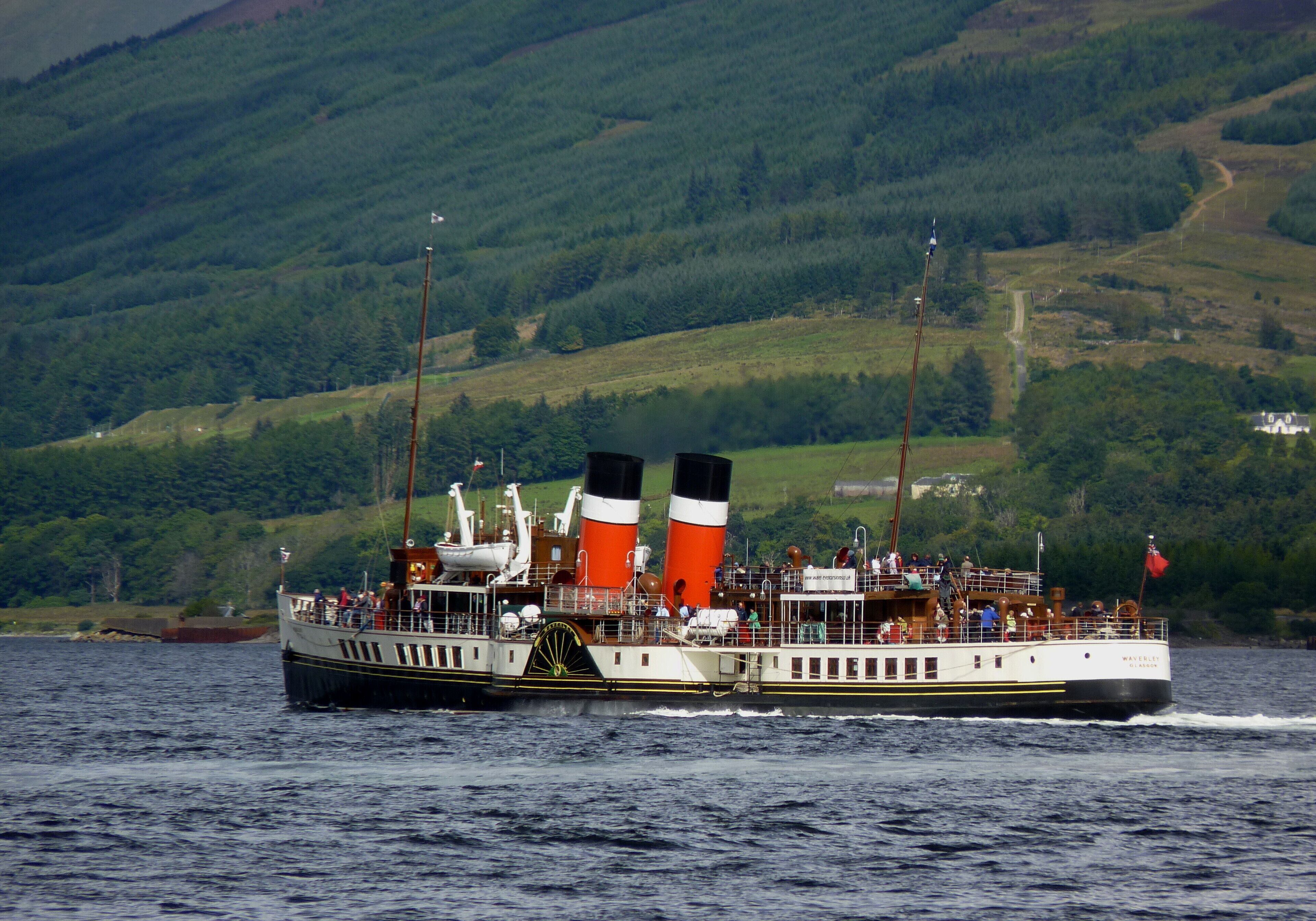 A steamboat with two prominent funnels on a body of water, surrounded by green hills.