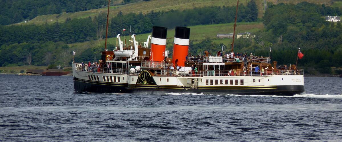 A steamboat with two prominent funnels on a body of water, surrounded by green hills.