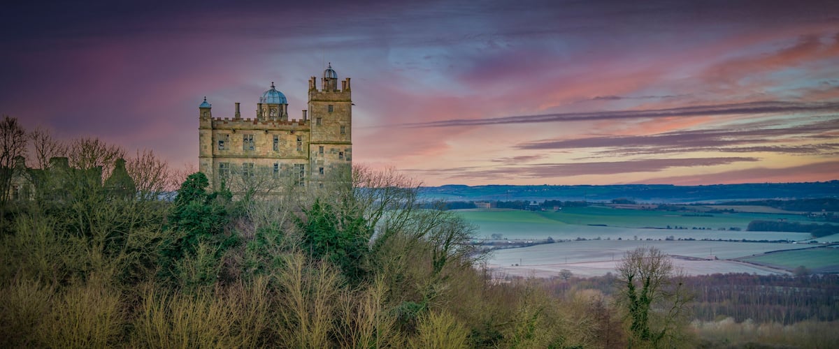 A castle surrounded by trees and fields with a colorful sky.