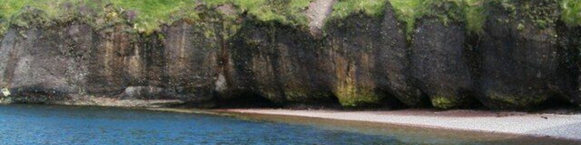 Cliff Undercutting at Roisinis. This picture, taken from a rocky outcrop, shows extensive undercutting of the cliffs. Cliff erosion is a major problem in the local area but this photograph depicting the erosion along with the blue sea and grass creeping down the cliff shows the damage as attractive rather than destructive.