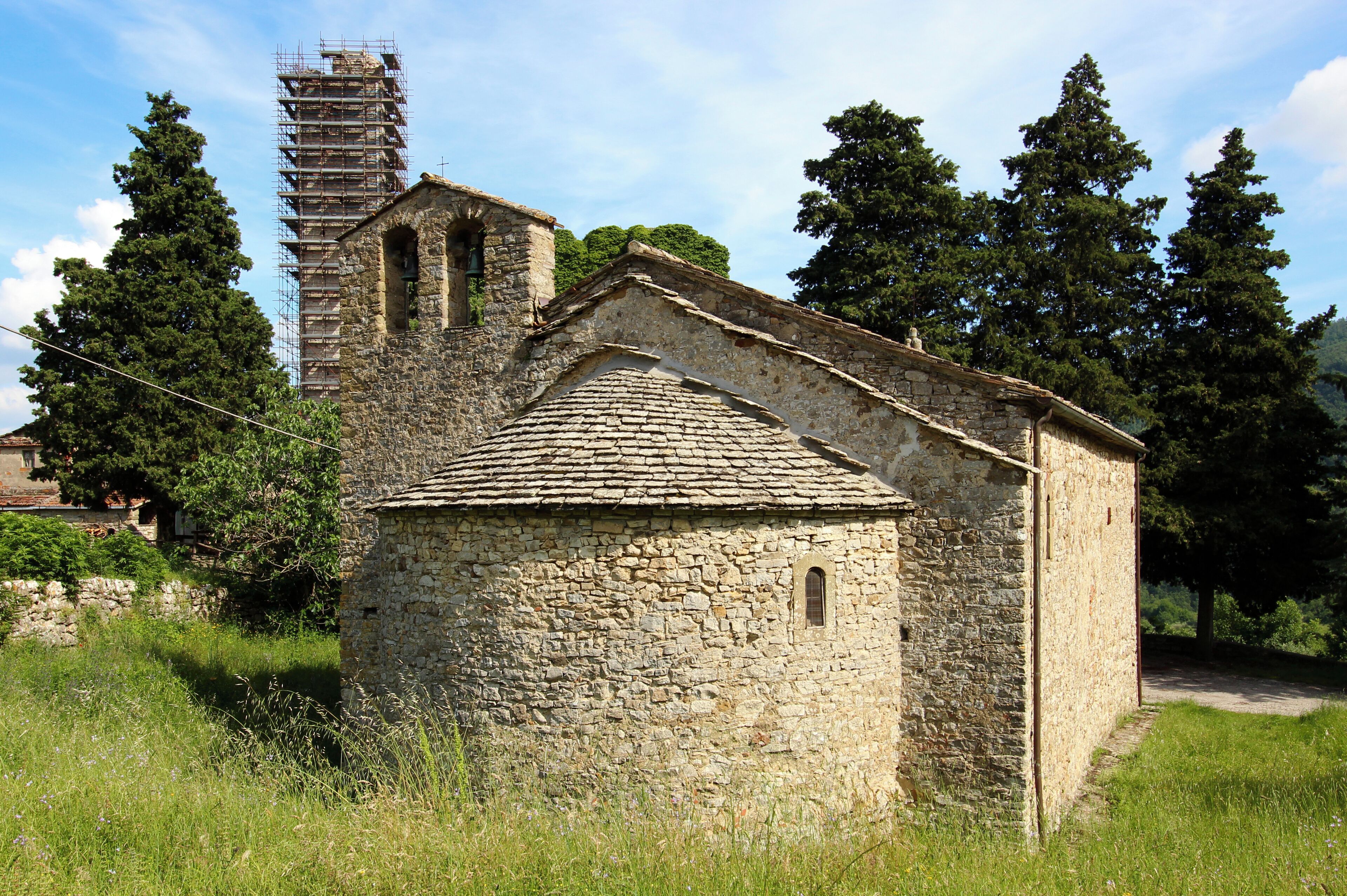 A stone church with a bell tower and scaffolding, surrounded by trees and grass.