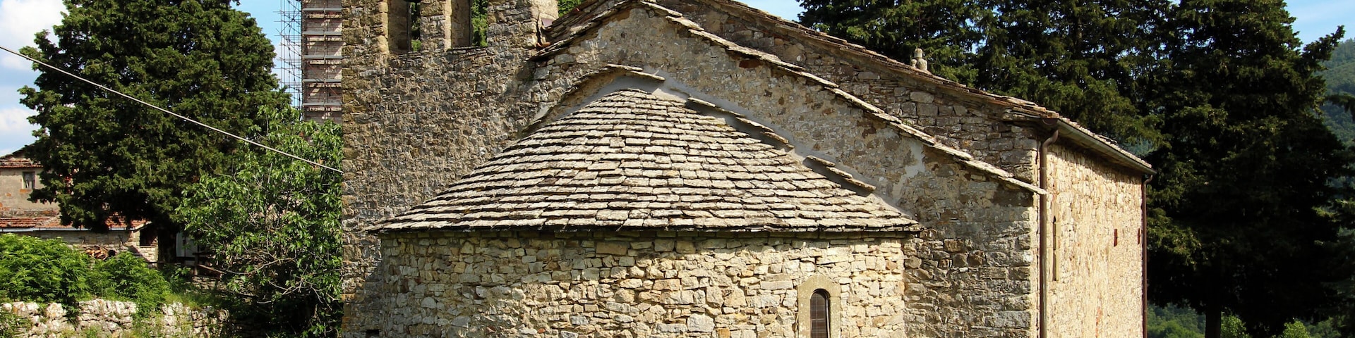 A stone church with a bell tower and scaffolding, surrounded by trees and grass.