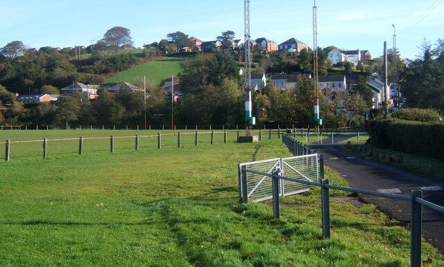 A grassy field with a soccer goal, a road, and houses on a hill in the background.