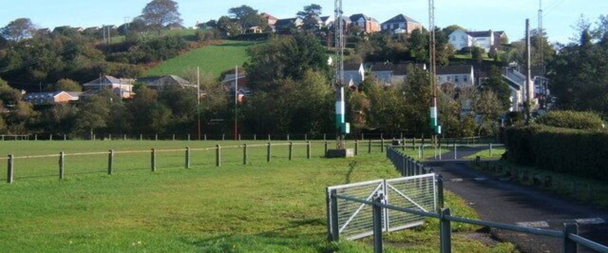 A grassy field with a soccer goal, a road, and houses on a hill in the background.