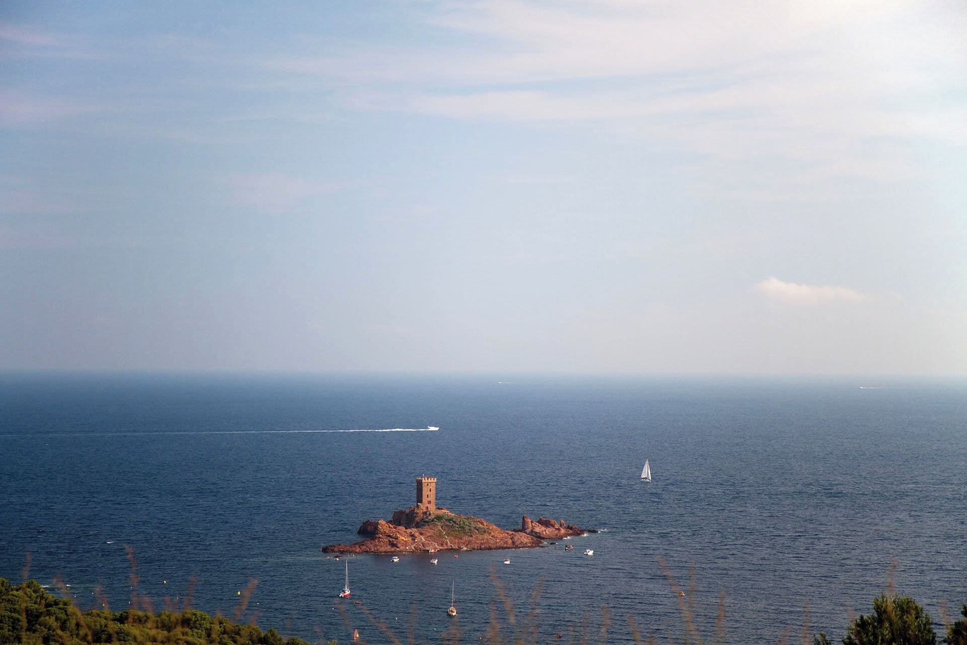 Paisaje costero con una pequeña isla rocosa que presenta una torre, rodeada de mar tranquilo y vegetación escasa.