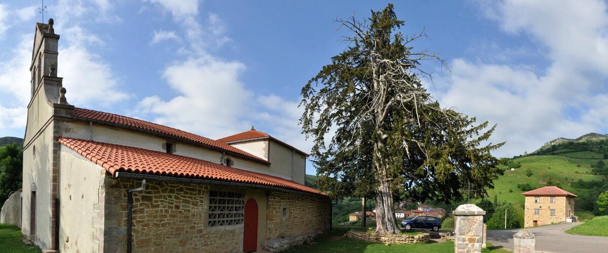 A rural landscape with a church, a stone building, a large tree, and rolling hills in the background.
