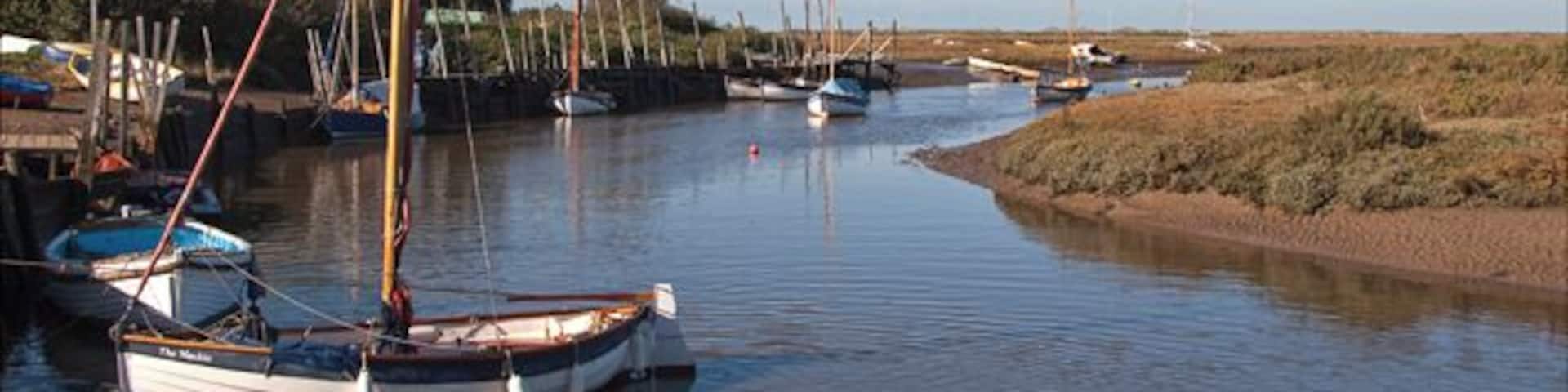 A calm river with boats docked on the left, a house, and open fields in the background.