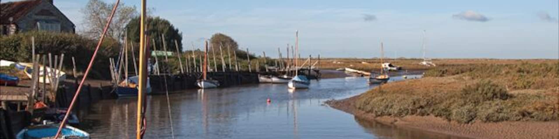 Un río tranquilo con barcas amarradas a la izquierda, una casa y campos abiertos al fondo.