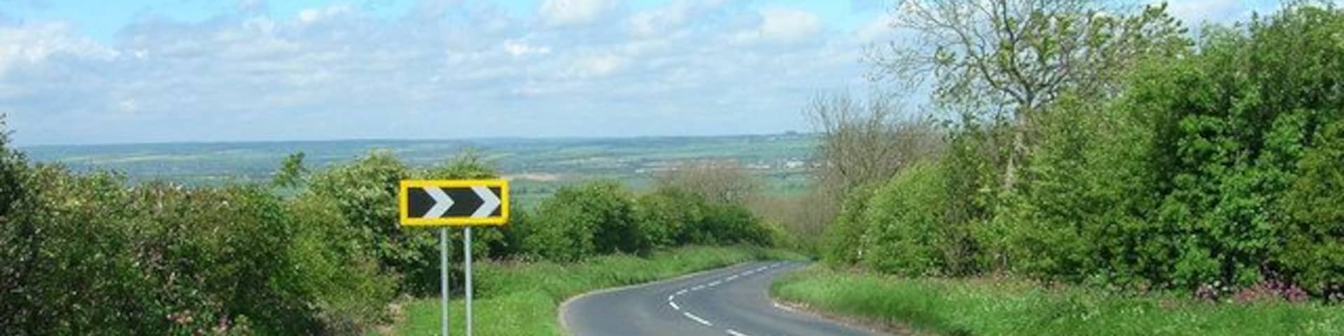 Uma estrada rural serpenteando para a direita, com uma placa de advertência amarela, cercada por vegetação e céu claro.
