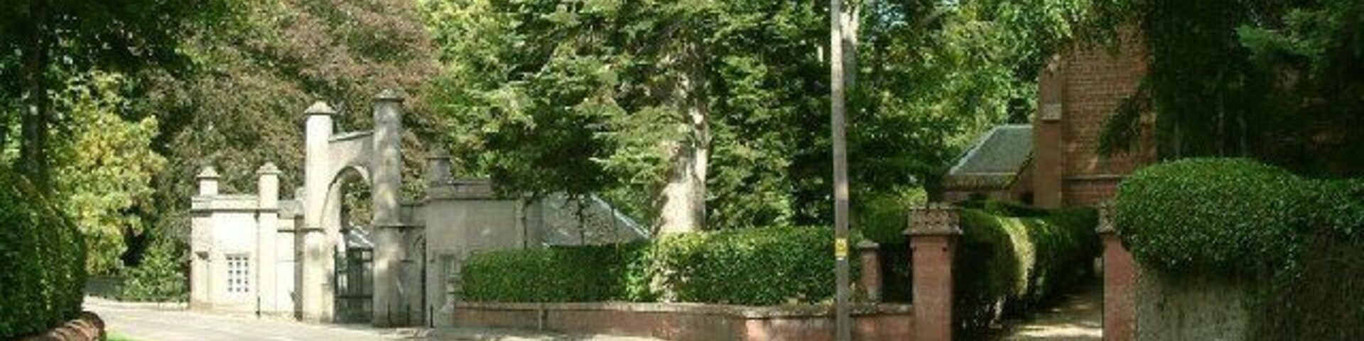 Uma rua residencial tranquila com portão fechado, muita vegetação e um edifício de tijolos.