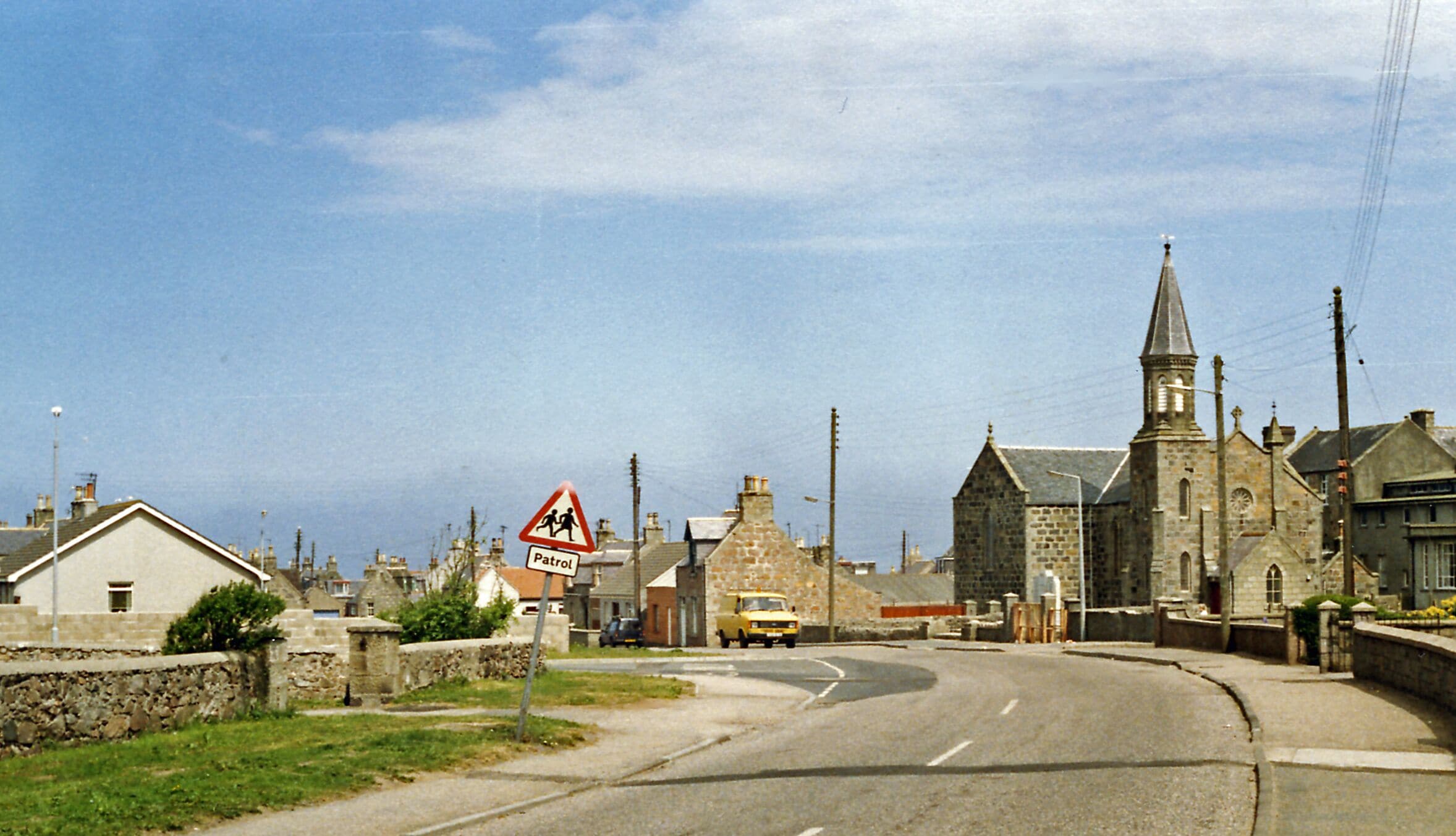 A rural road with a church, a 'Patrol' sign, and a 'School Crossing' sign.
