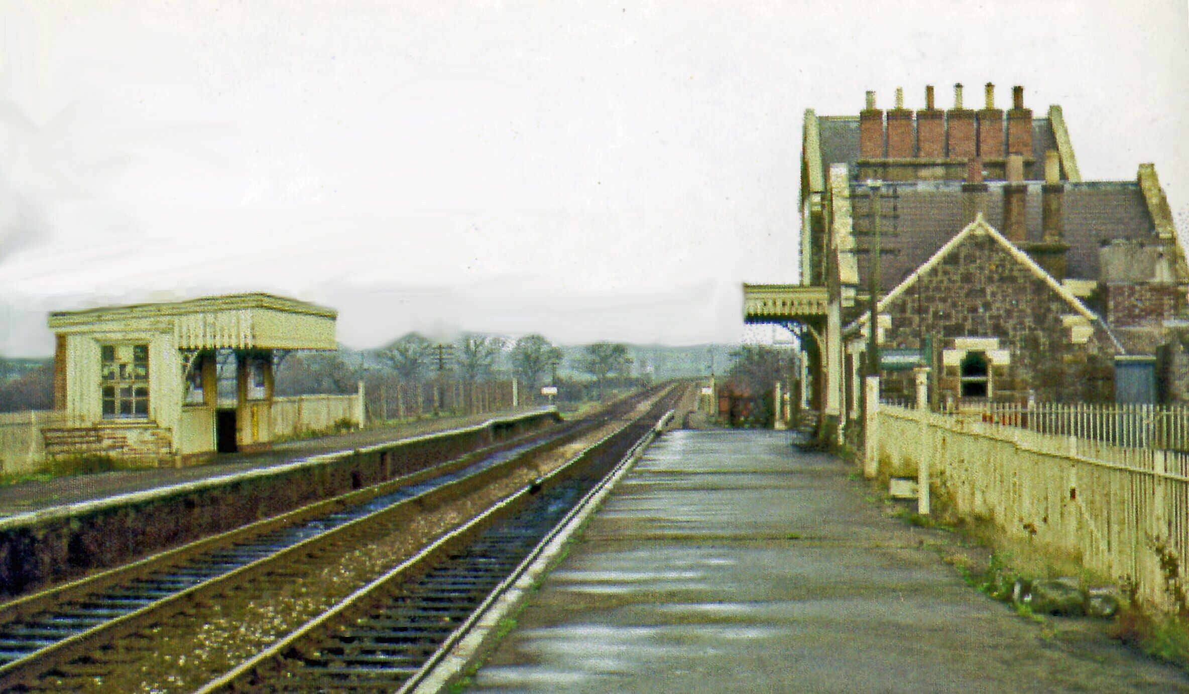 A railway station with tracks, a small waiting area, and a large stone building.
