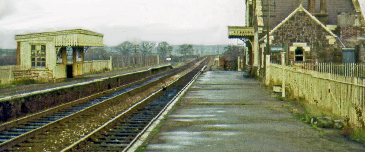 A railway station with tracks, a small waiting area, and a large stone building.