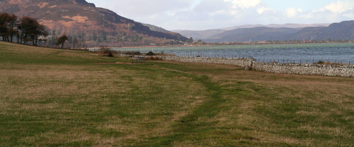 A grassy field with a body of water, a stone wall, and hills in the background.