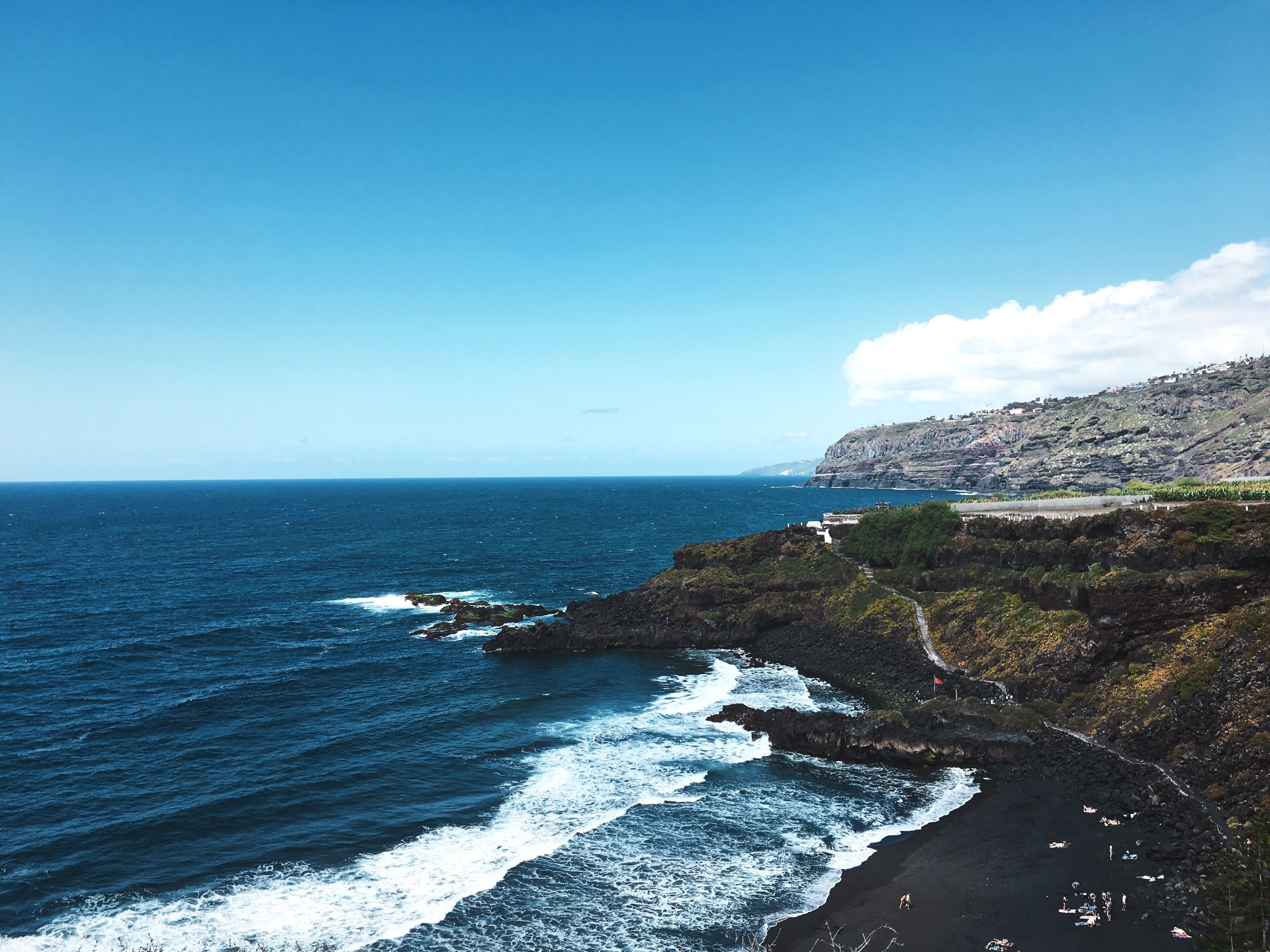 A coastal landscape with a rocky shoreline, waves crashing onto the beach, and a clear blue sky.