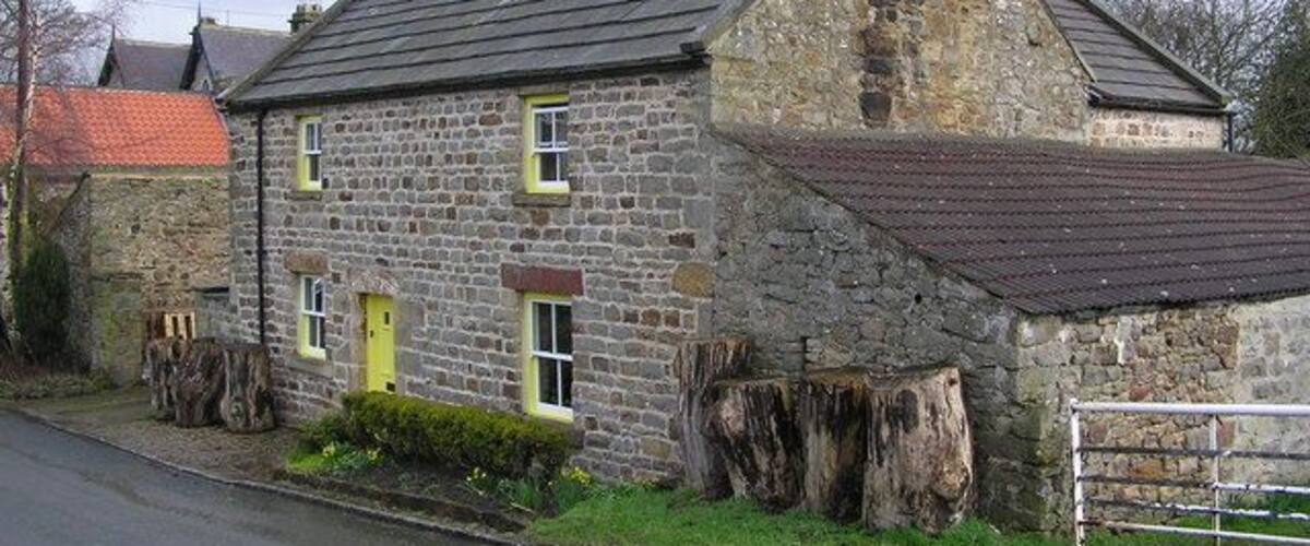 A stone house with a sloped roof, a yellow door, and a large tree stump in front.