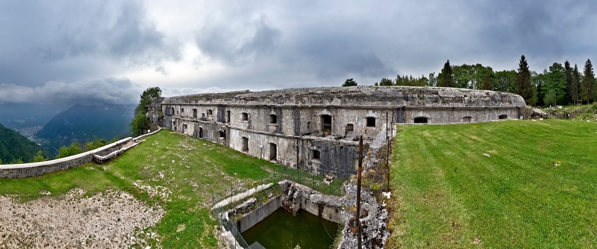 A historic stone building with a grassy courtyard and a small pond, surrounded by mountains and trees under a cloudy sky.