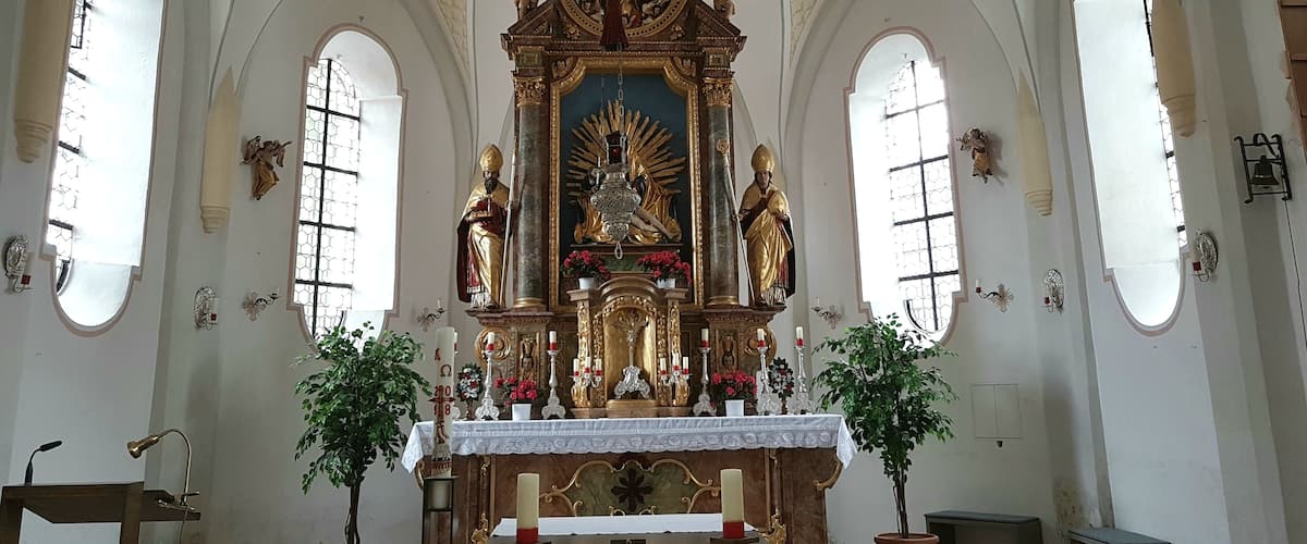 An ornate church interior with a central altar, arched windows, and religious statues.