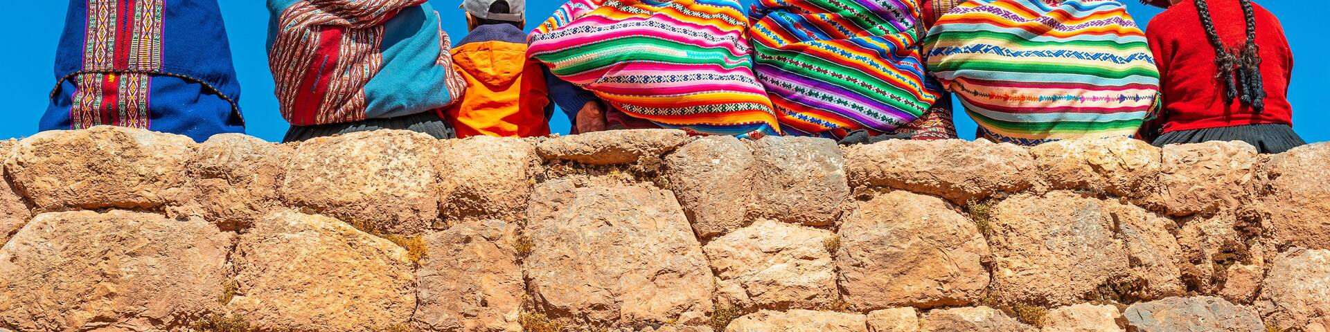 Panoramic photograph of Quechua indigenous women in traditional clothing with a boy sitting on an ancient Inca wall in Chinchero, Cusco Province, Peru.