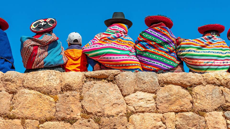 Panoramic photograph of Quechua indigenous women in traditional clothing with a boy sitting on an ancient Inca wall in Chinchero, Cusco Province, Peru.