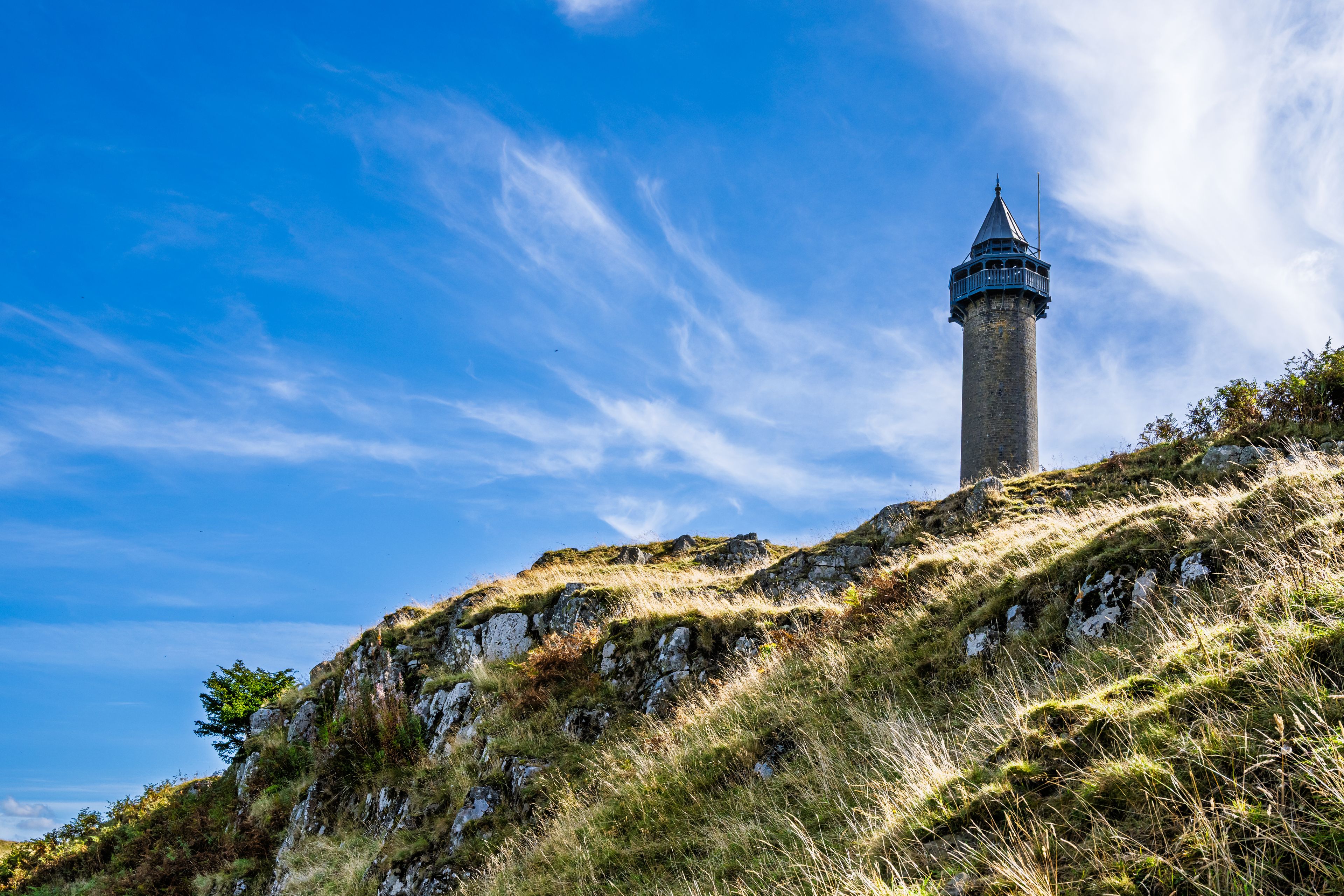 Una torre alta con techo puntiagudo, rodeada de colinas cubiertas de hierba y un cielo azul despejado.