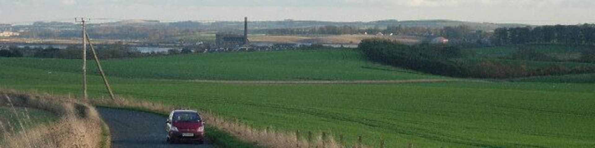 Minor road linking Balmullo and Leuchars. The view is east south east along the road from the north of the grid box. The chimney of the paper mill at Guardbridge is visible in the distance.