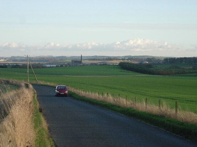 A rural road with a red car driving on it, surrounded by green fields and distant hills.
