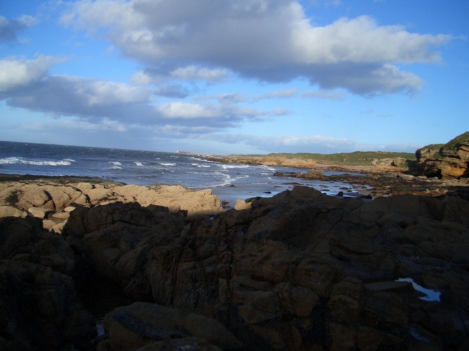 A rocky coastline with waves crashing against the shore under a partly cloudy sky.
