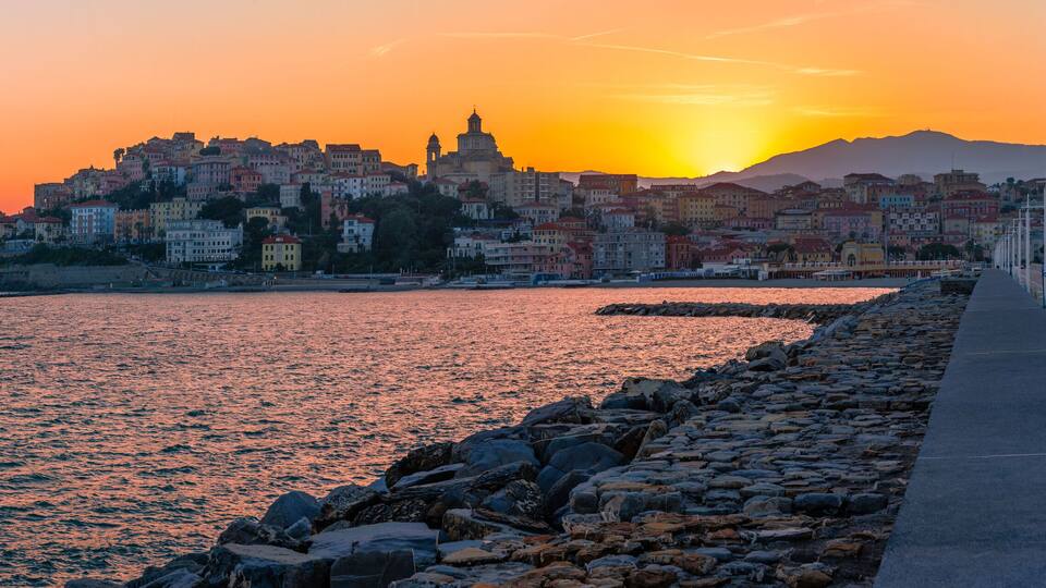 A coastal cityscape at sunset with a prominent church, a stone pier, and colorful buildings.