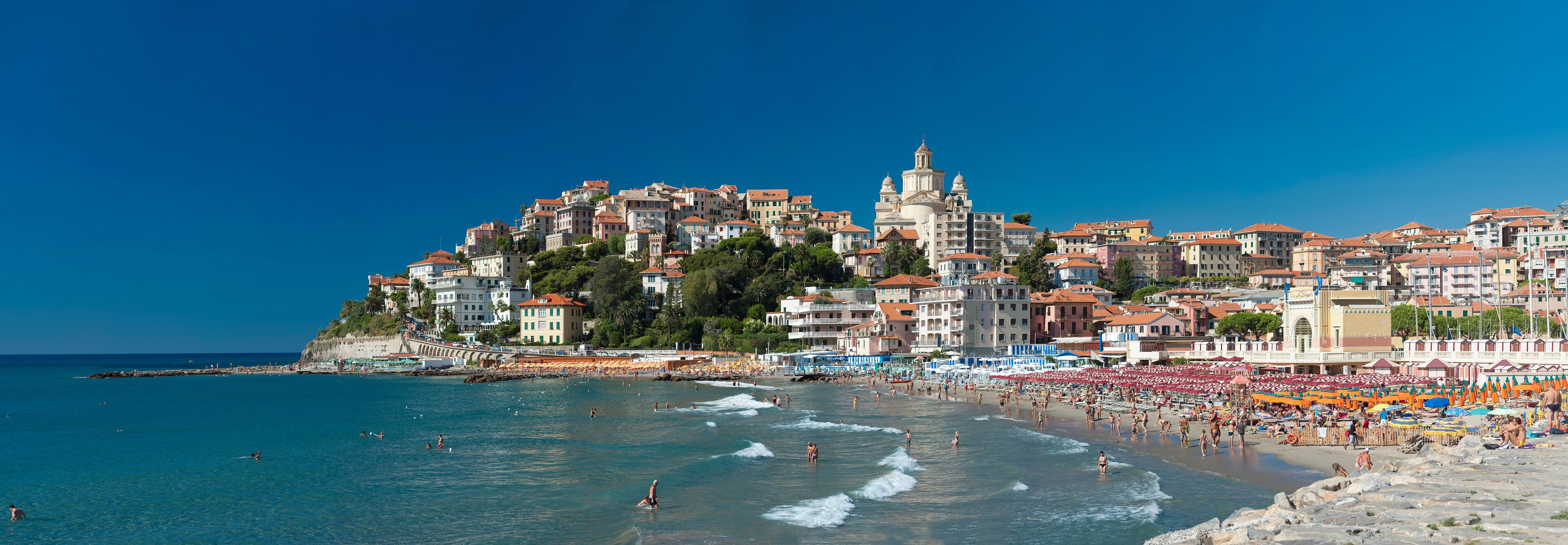 A coastal town with a beach, people swimming, and a prominent church on a hill.