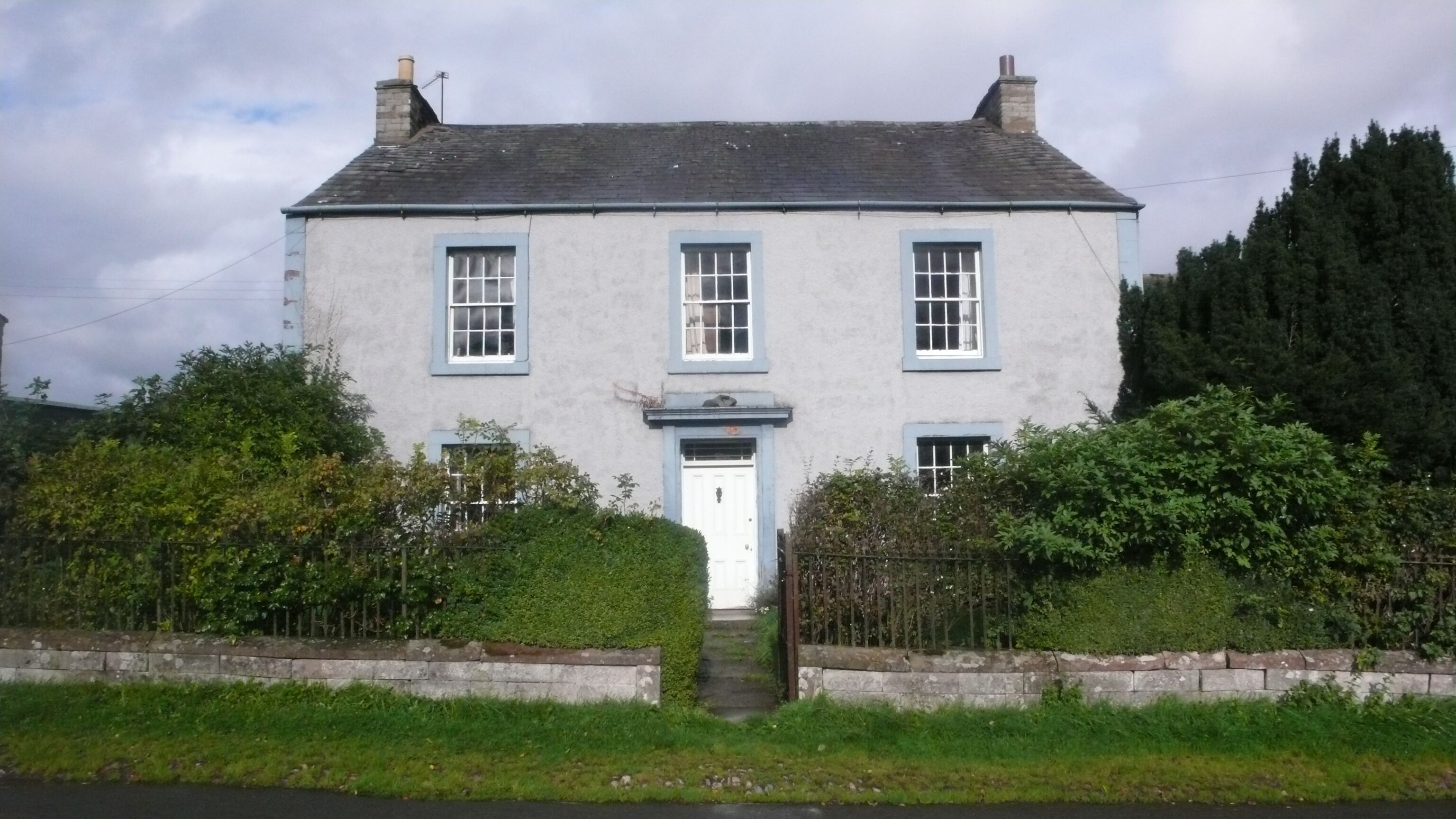 A two-story house with a white door and blue trim, surrounded by greenery and a stone wall.