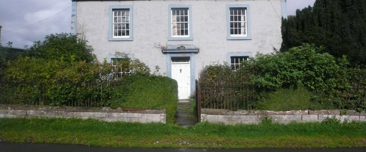 A two-story house with a white door and blue trim, surrounded by greenery and a stone wall.