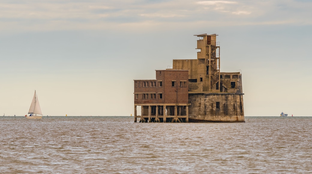 Un edificio ruinoso sobre pilotes en el agua, con un velero al fondo.