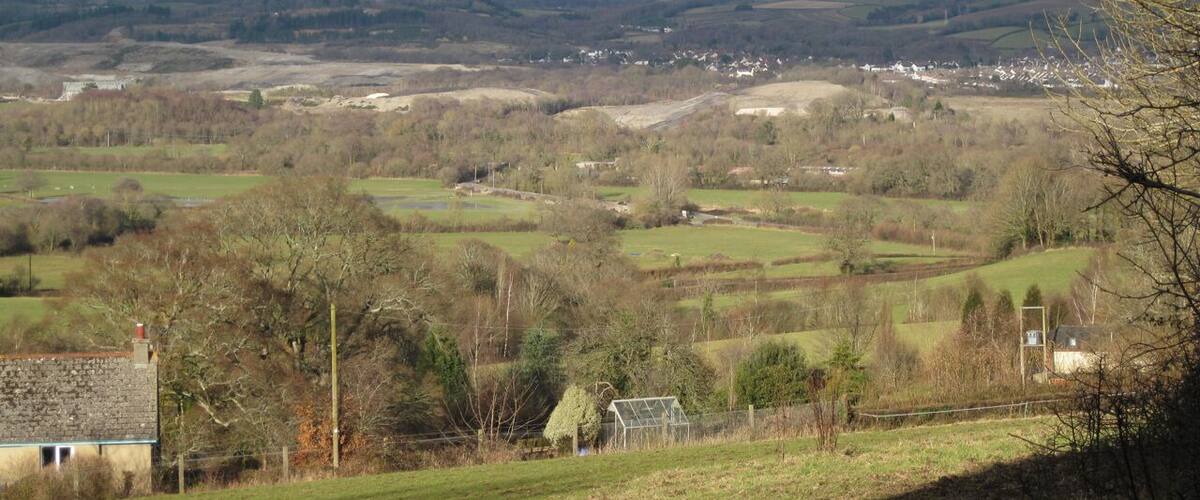 View of the Bovey Basin from Whitehill Road (3/3) Looking northeast over Higher Loady Park from below the cottages 1729963. In the middle distance are floodmeadows on the west bank of the Teign, crossed by the racetrack which is the old Exeter Road. In the centre of the photo, hidden by trees, is Teign Bridge. Left of centre is a mound of clay waste being formed on the east side of Denistone Quarry SX8574, with a wooded mound of old waste appearing to its left SX8573. Clay waste stretches away to the north. Right of centre the Broadway clay pit is being filled, with a big shaped mound to its right. Beyond it at some distance is Higher Sandygate SX8674, mostly. Between the mounds in the centre of the photo is the narrow wooded cleft which marks the course of the Abb Brook or Abbrook which, in its upper reaches, is the Ugbrooke Stream SX8676.