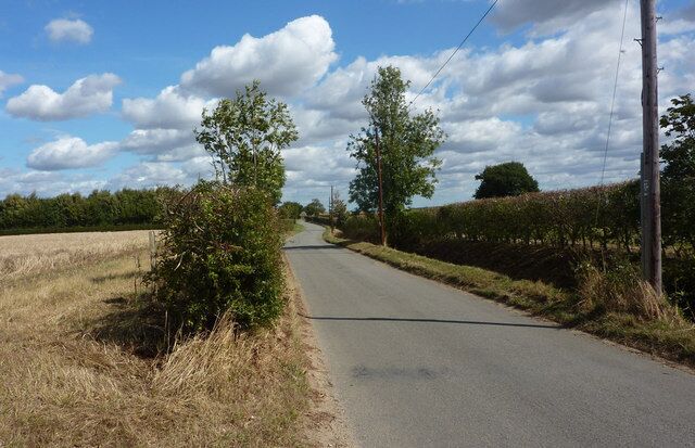 A rural road flanked by fields and trees under a partly cloudy sky.