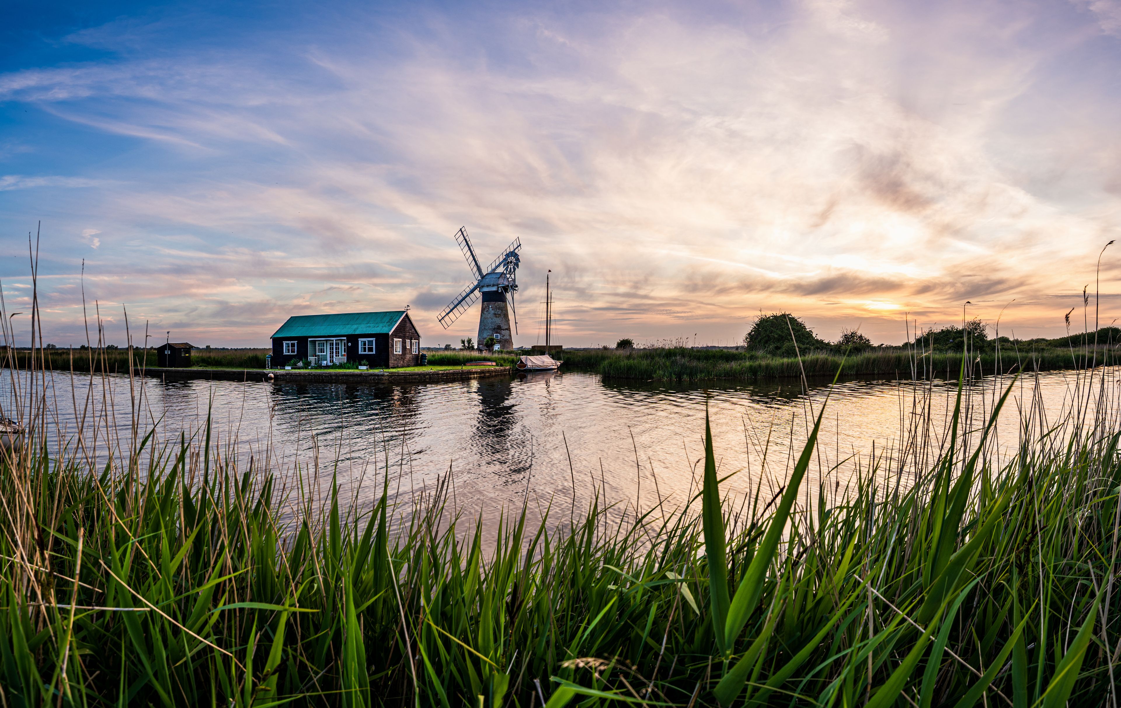 A windmill, a house, and a body of water with tall grass in the foreground.