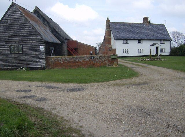 A gravel path leading to a white house with a black roof, a wooden barn, and a brick wall.