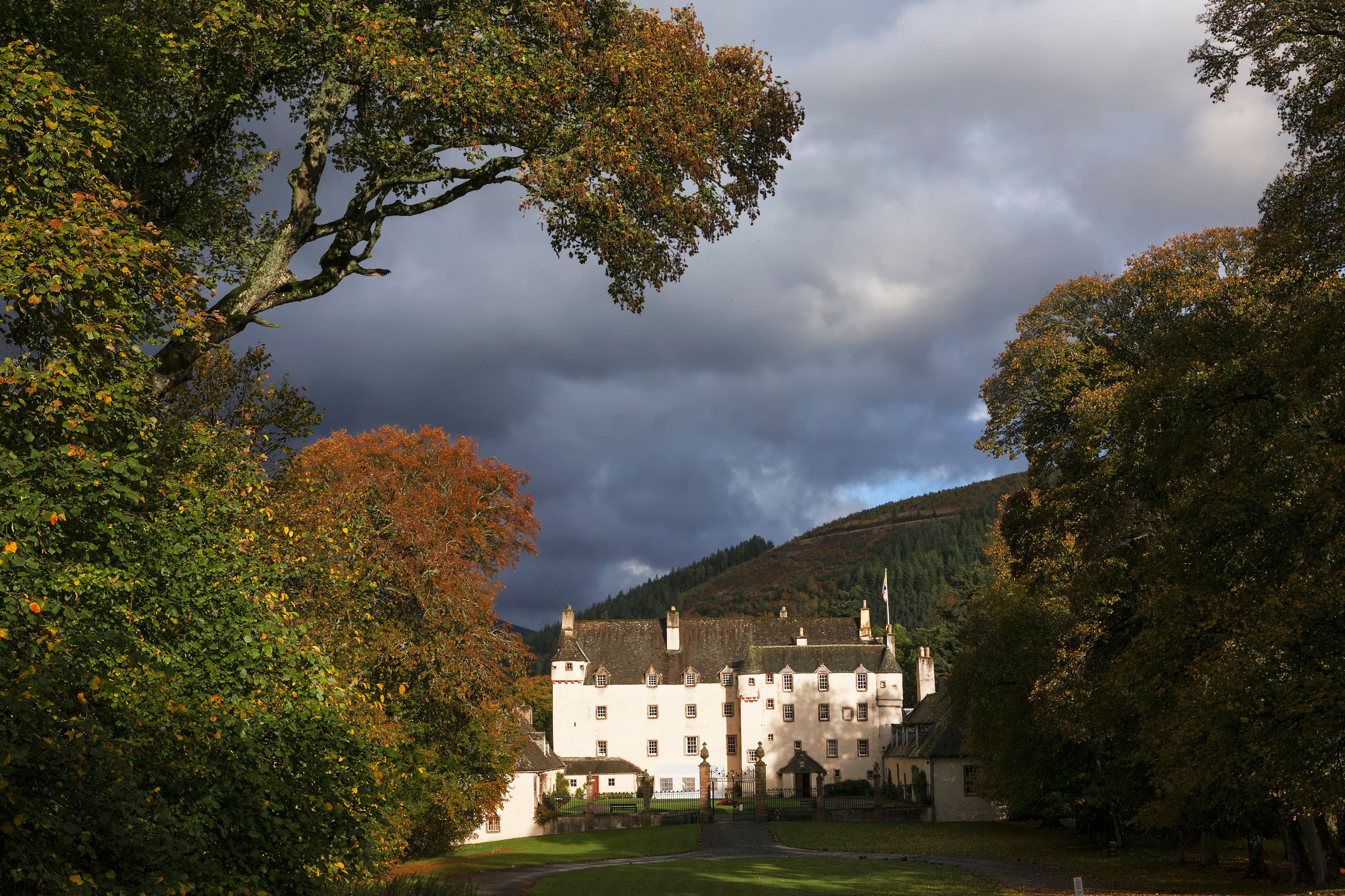 A large white building with multiple windows and a flag, surrounded by trees with autumn leaves, under a cloudy sky.