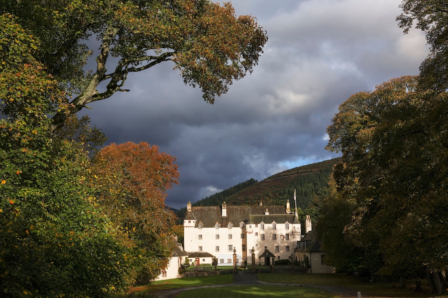 A large white building with multiple windows and a flag, surrounded by trees with autumn leaves, under a cloudy sky.