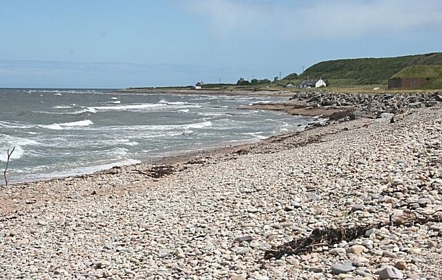 A rocky beach with waves, a grassy hill, and a few buildings in the distance.