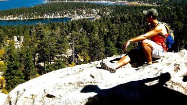 Una persona sentada sobre un afloramiento rocoso con vistas a un pueblo costero rodeado de bosques densos y montañas.