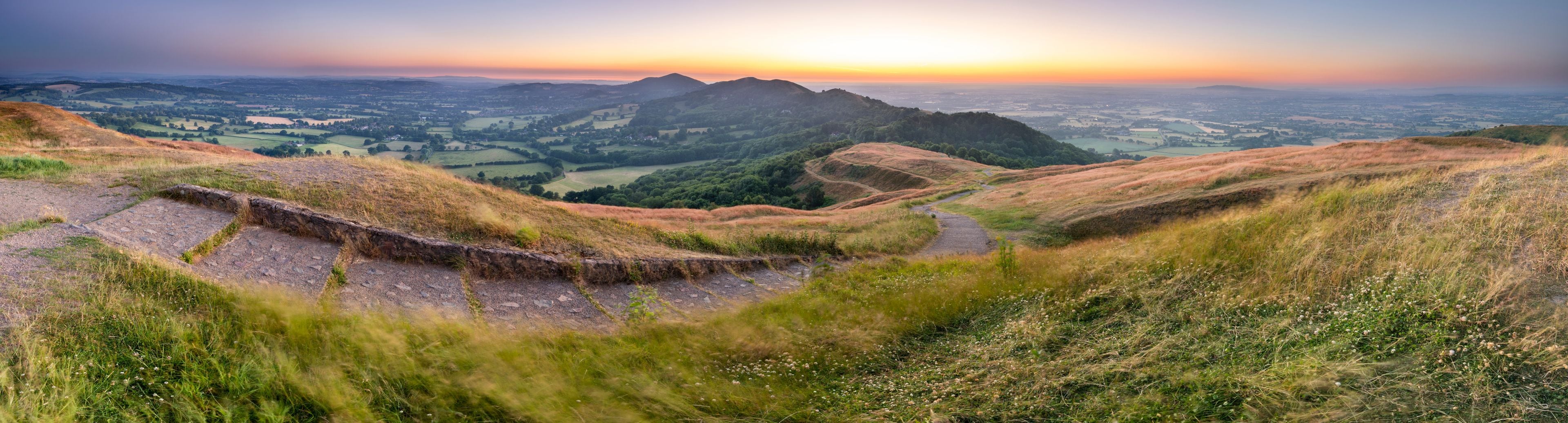 Un paisaje montañoso con un atardecer, un camino sinuoso y campos lejanos.