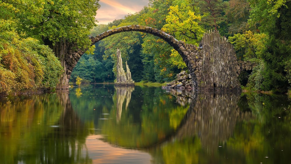 Eine steinernen Bogenbrücke überspannt einen ruhigen Fluss, umgeben von üppigem Grün.